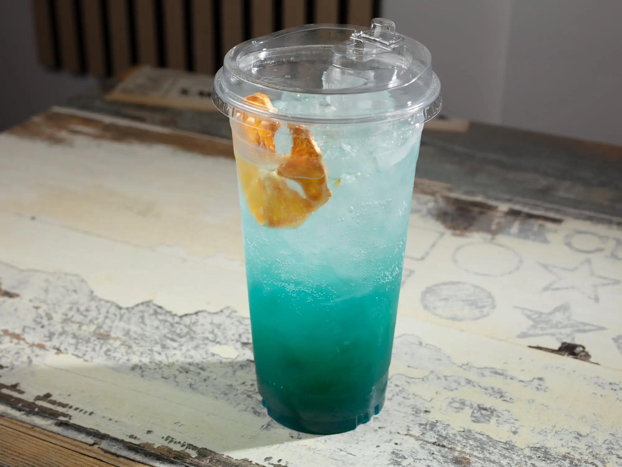 A tall clear cup with a domed lid, filled with bright blue iced soda and a dried citrus wheel floating near the top
