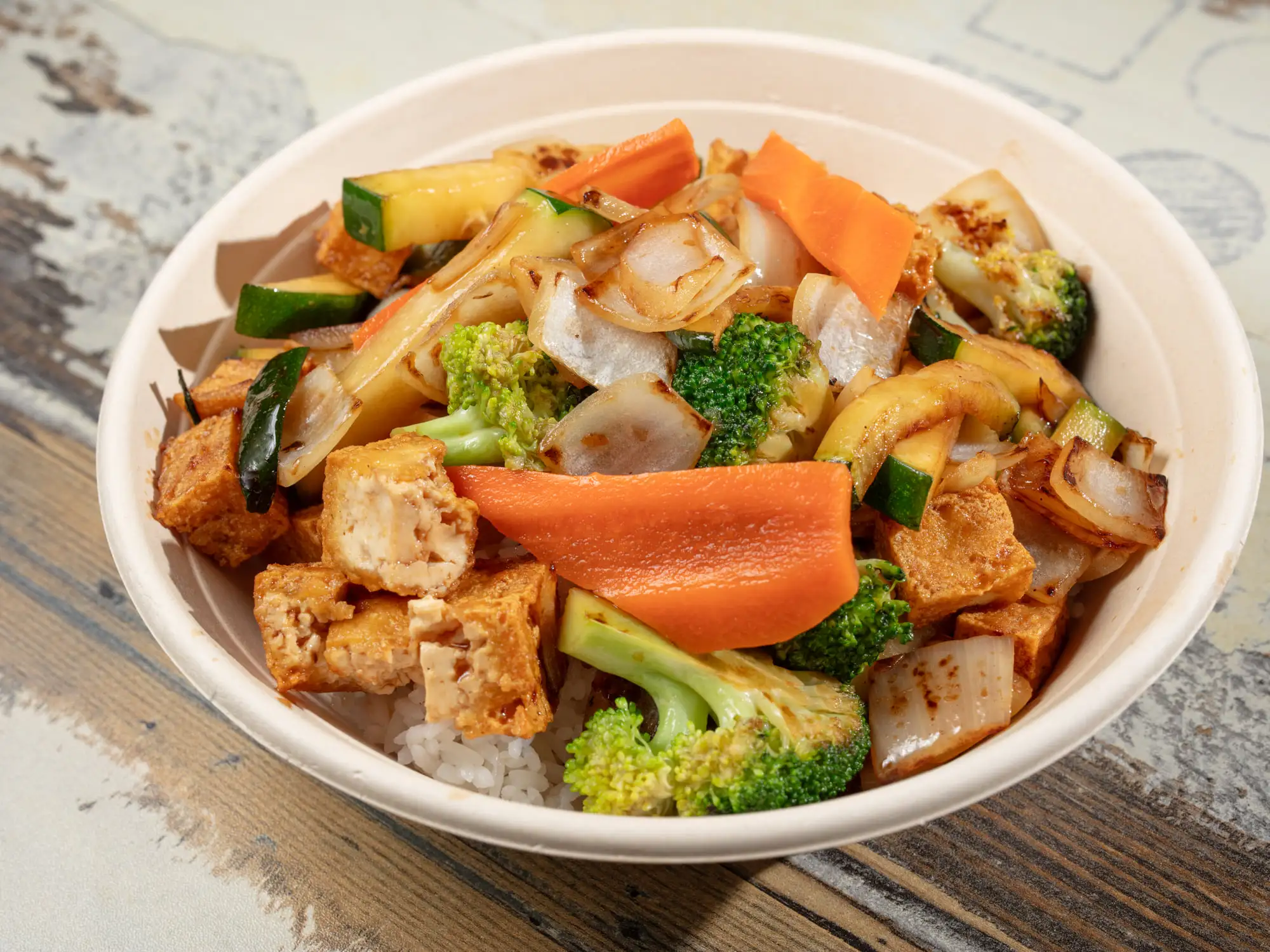 Tofu with zucchini, carrot, onion and broccoli over white rice in a white bowl, glossy with a brown house sauce