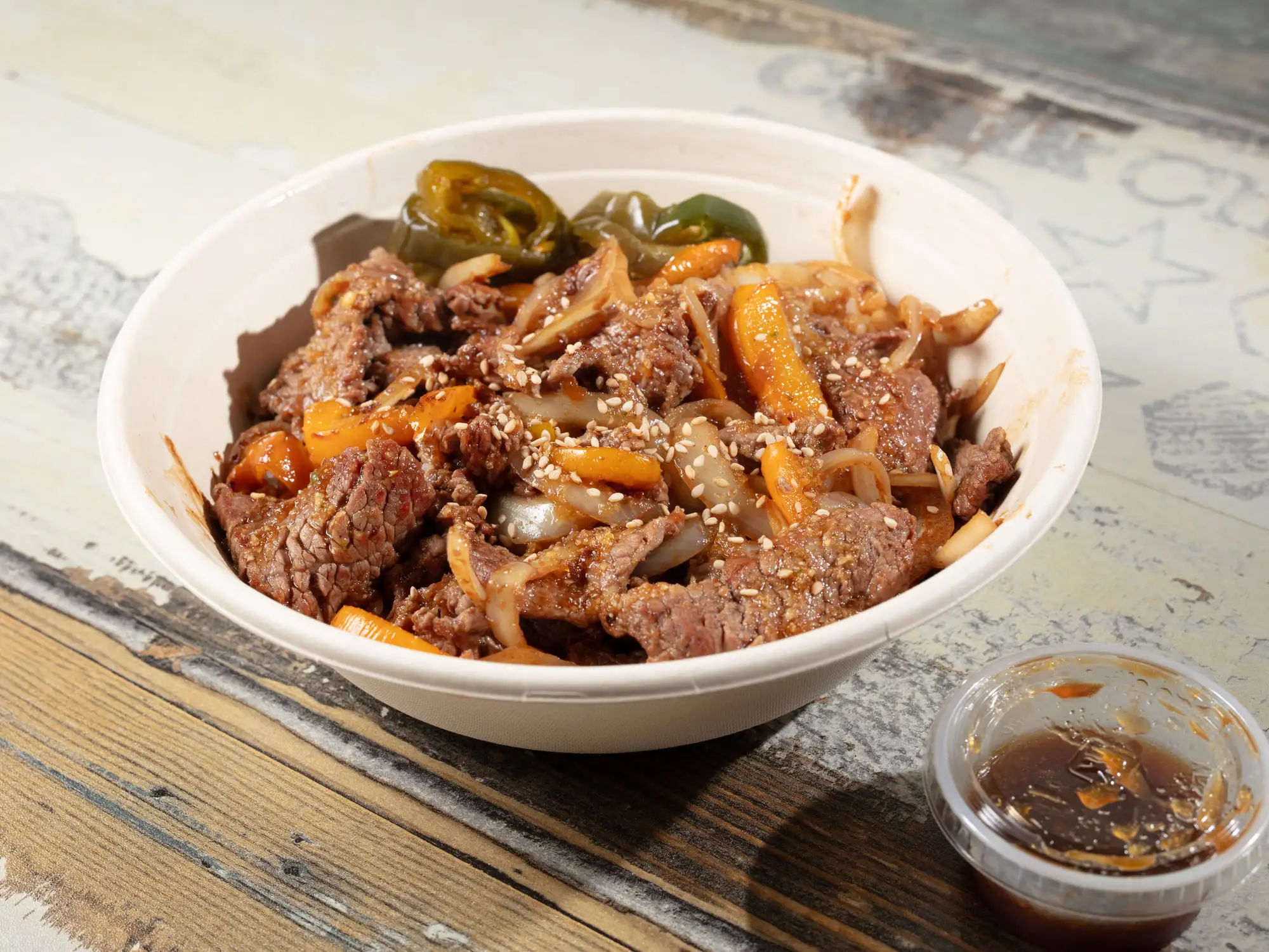 Thin-sliced marinated bulgogi beef with onion and carrot in a white bowl, sesame seeds and blistered green jalapenos on top, a lidded cup of dark sauce beside it