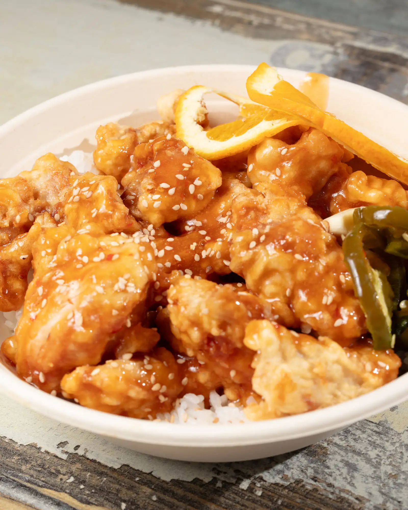 Orange-glazed fried chicken over white rice in a white bowl, finished with sesame seeds, a twist of orange zest and blistered green peppers