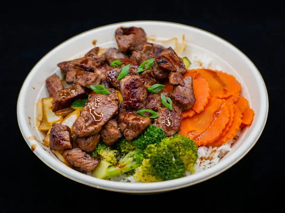 Seared steak with green onion over white rice in a white bowl, with crinkle-cut carrots, broccoli, onion and zucchini