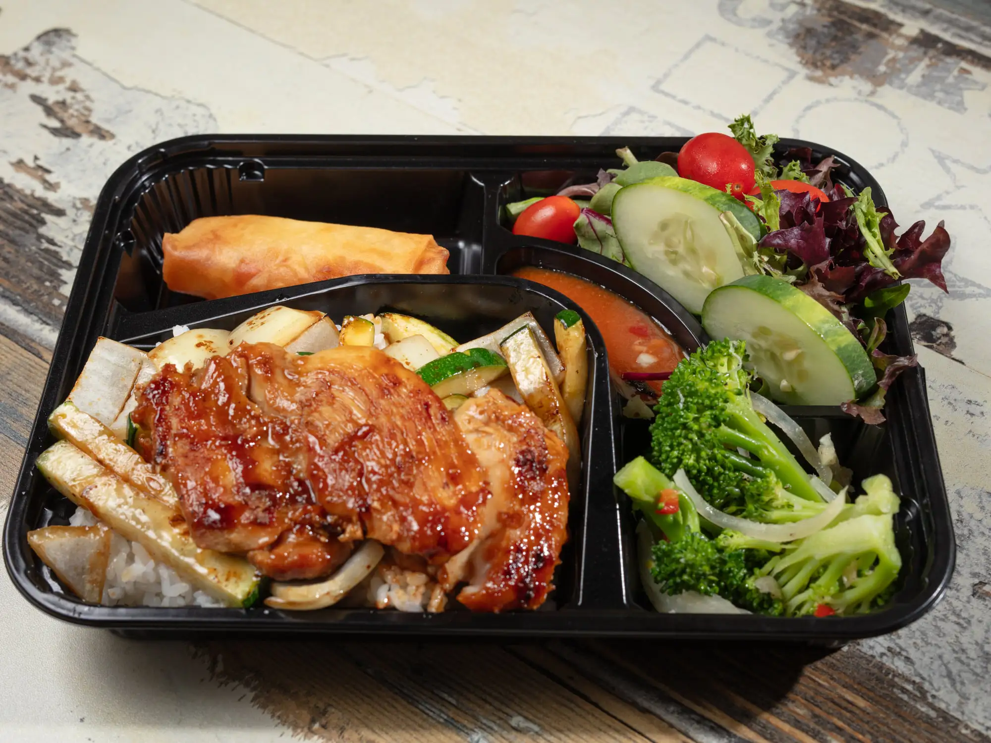 A black bento tray holding glazed chicken over griddled onion and zucchini, a spring roll, and a salad of mixed leaves, cucumber, tomato and broccoli
