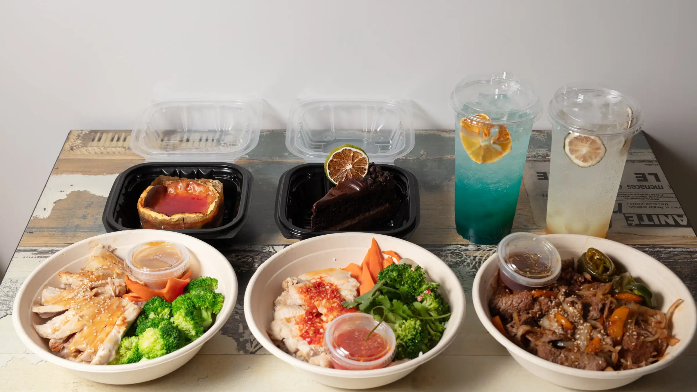 Three hibachi bowls with chicken, tofu and bulgogi over rice with broccoli and carrots, beside two house drinks and a slice of chocolate cake