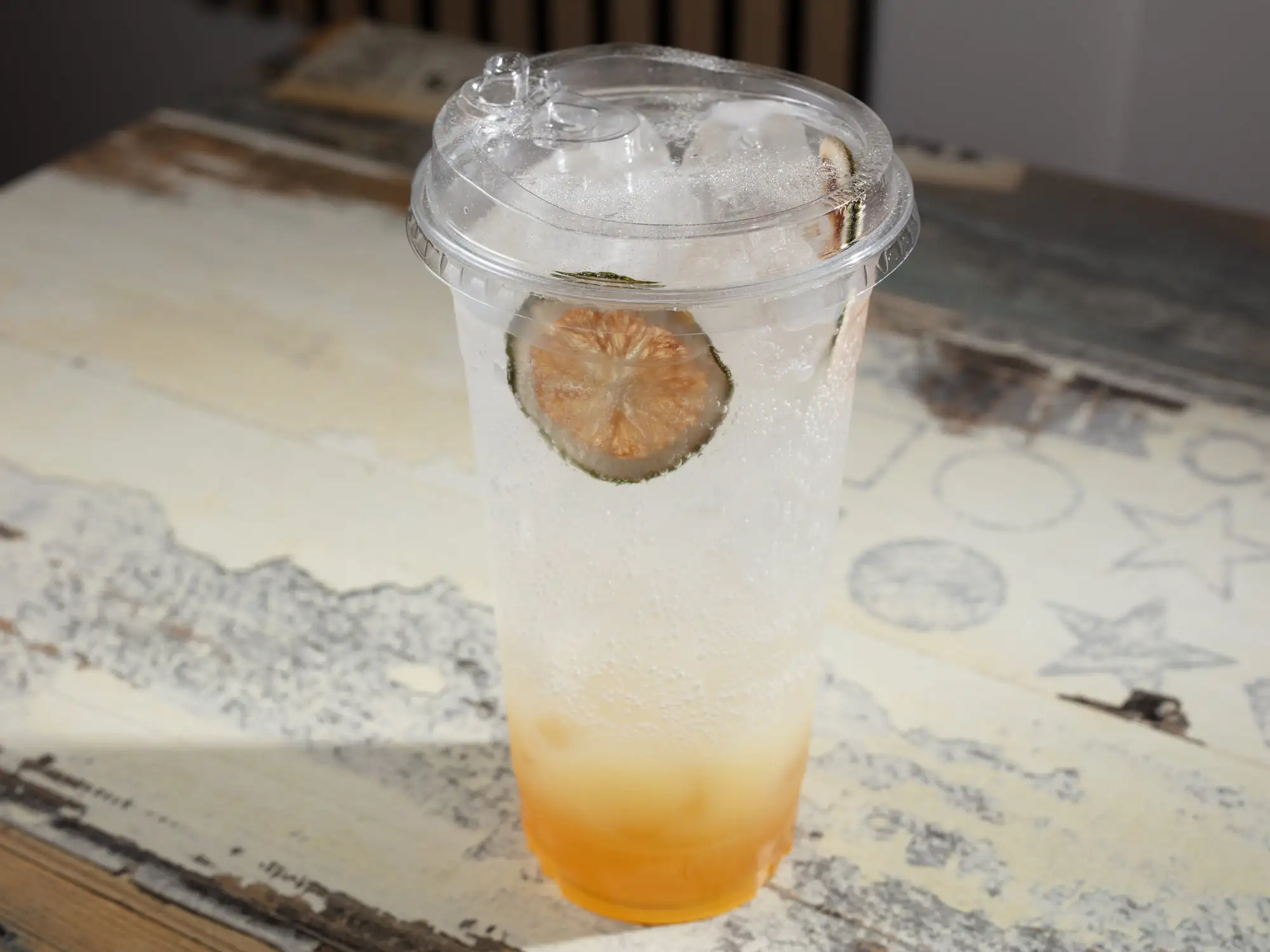 A tall clear cup with a domed lid, pale sparkling citrus soda over a deeper amber base, packed with ice, a dried lime wheel on top
