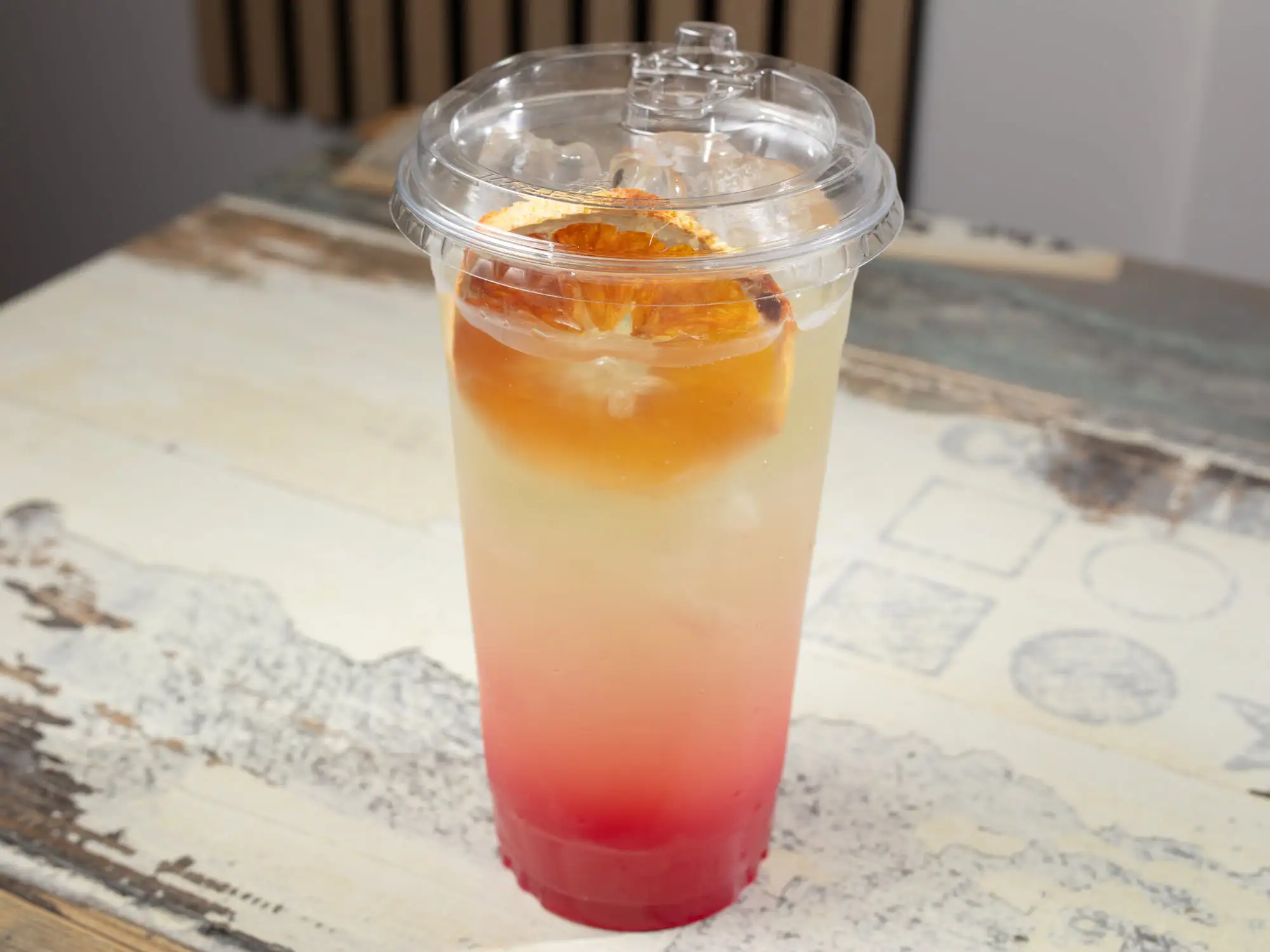 A tall clear cup with a domed lid, the drink graded from deep red at the base through orange to pale yellow, over ice, with a dried orange wheel on top