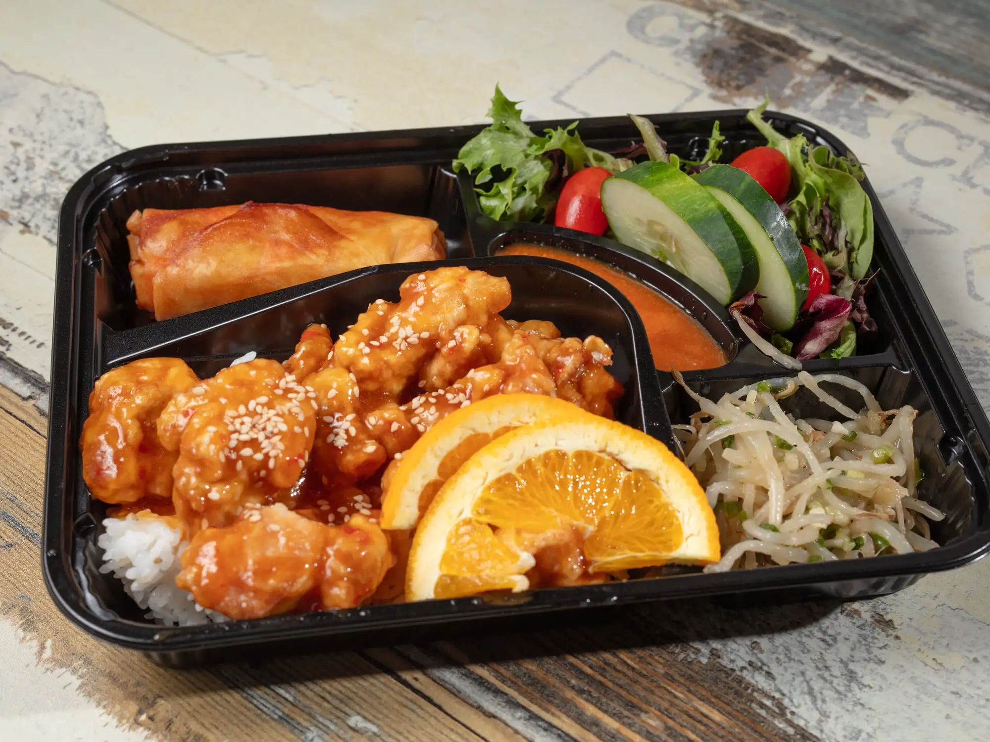 A black bento tray holding orange-glazed fried chicken over rice with two orange slices, a spring roll, bean sprouts, and a side salad with cucumber and tomato
