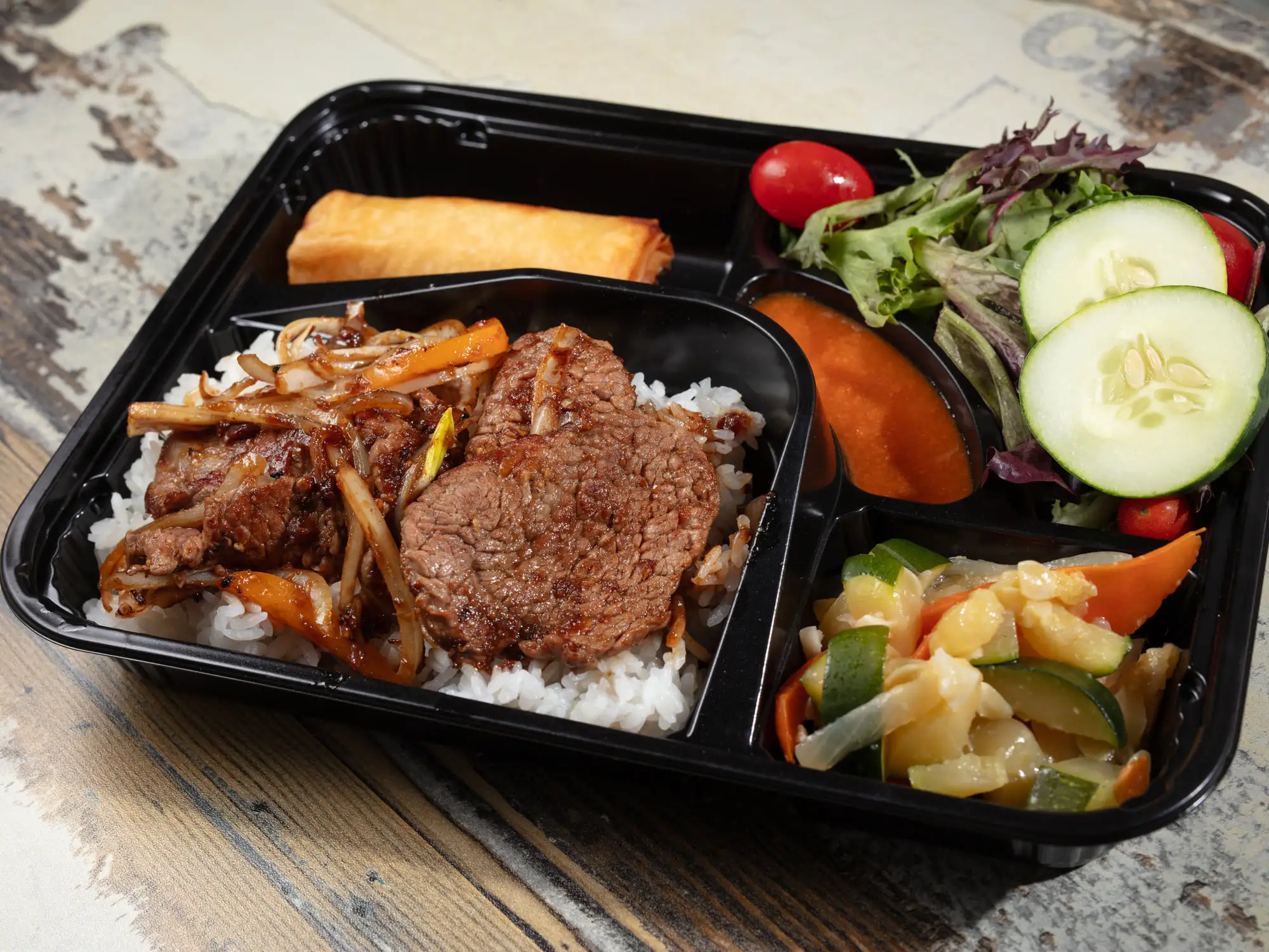 A black bento tray holding sliced beef with onion and carrot over white rice, a spring roll, griddled vegetables, and a salad of mixed leaves, cucumber, tomato and carrot