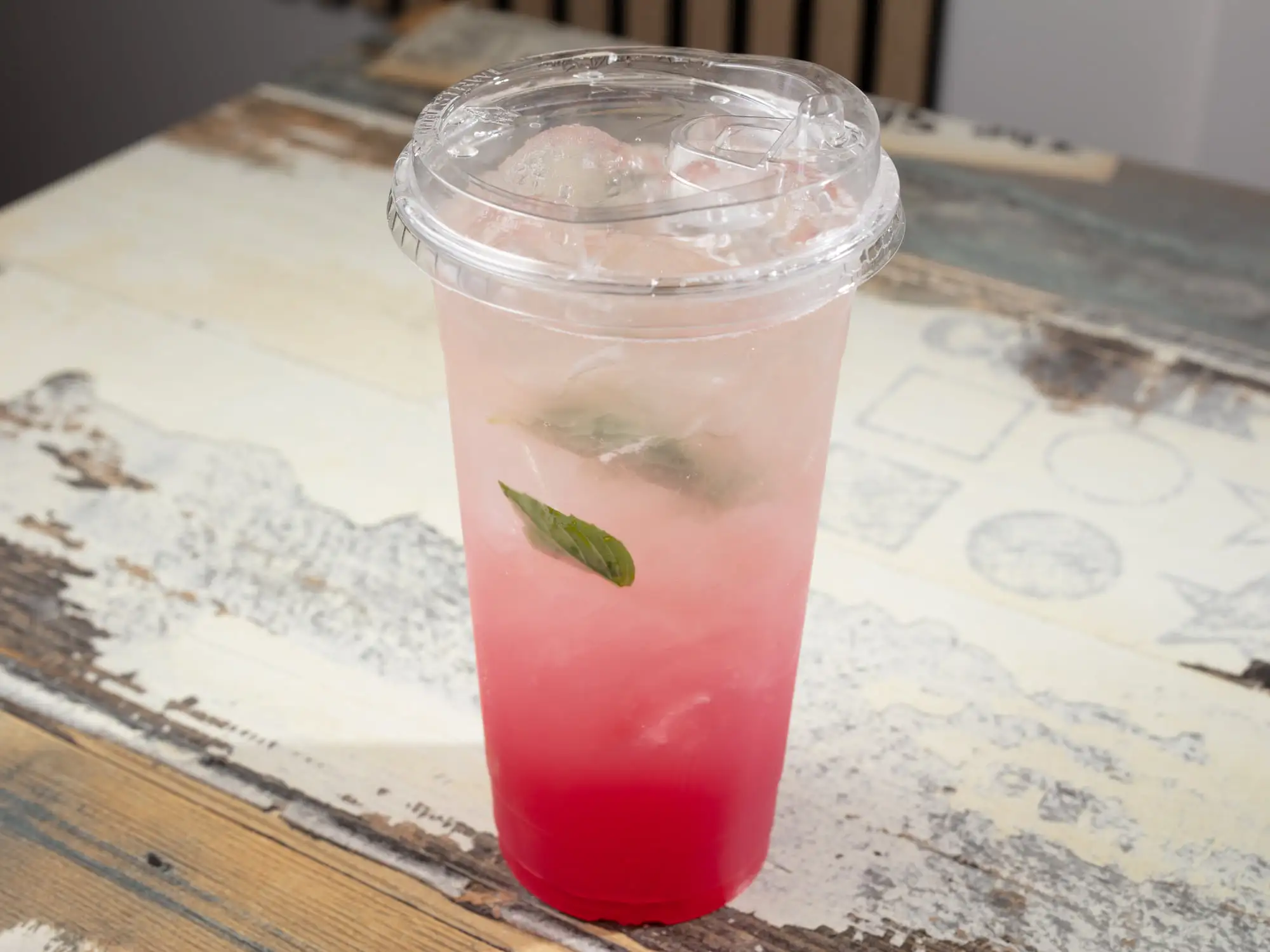 A tall clear cup with a domed lid, pink drink deepening to red at the base, over ice, with a fresh basil leaf floating on top