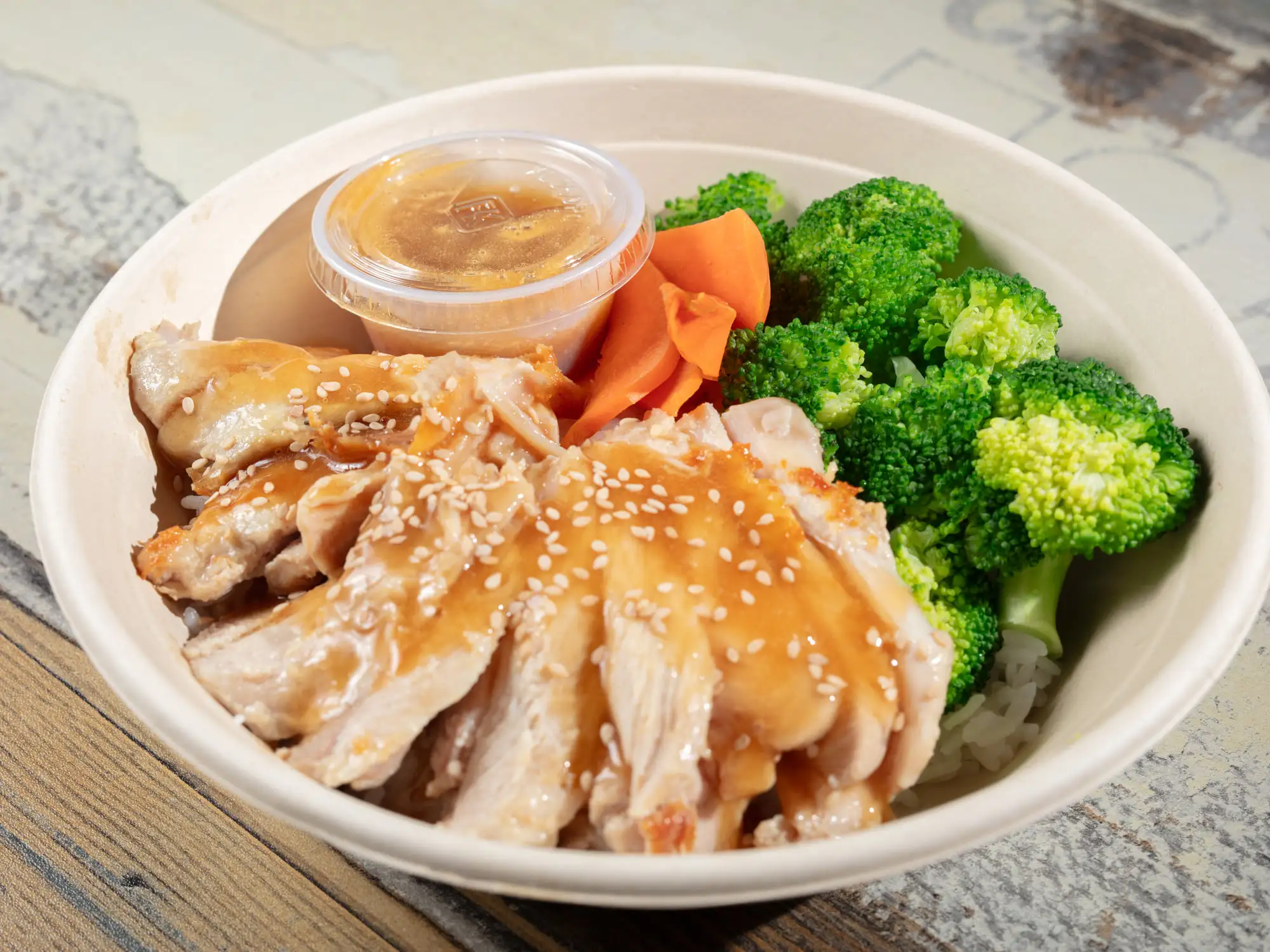 Sliced teriyaki-glazed chicken fanned over white rice in a white bowl, scattered with sesame seeds, with steamed broccoli and a curl of carrot alongside and a lidded cup of sauce behind